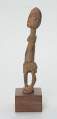 Drewniana figurka siedzącej postaci - Ujęcie z lewego boku; figurka przedstawiająca postać ludzką w pozycji siedzącej. Części ciała i rysy twarzy wydobyte za pomocą płytkich nacięć siekierki.