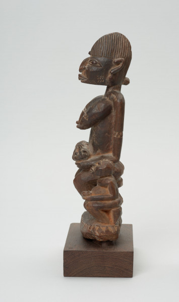 rzeźba - Ujęcie z boku; Figura - kobieta z dziećmi. Siedząca bokiem kobieta trzyma na rękach niemowle, które opiera na je na kolanach. Przy niej siedzi małe dziecko.