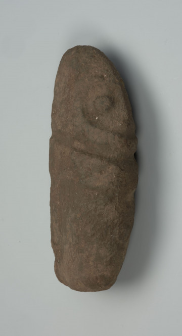 Kamienna figura antropomorficzna - Ujęcie z lewej strony; obiekt leżący; kamienna figura owalna, podłużna, rzeźbiona płytkimi uderzeniami, z zaznaczonymi oczami i wąskim nosem.