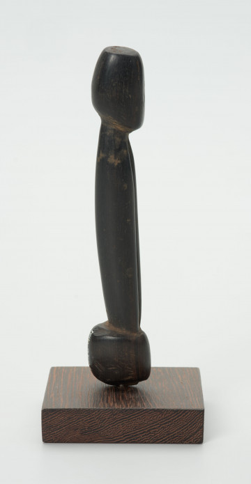 Drewniana figurka postaci z długim tułowiem - Ujęcie z prawej strony; drewniana figurka postaci ludzkiej przedstawionej schematycznie. Całość wygładzona, zarysowana głowa i długi tułów.
