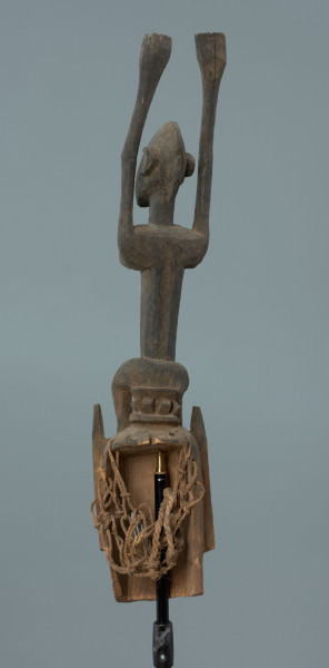 drewniana maska - Ujęcie z tyłu; Drewniana, rzeźbiona maska. Figura postaci ludzkiej z rękoma wzniesionymi do góry. Głowa jajowata, spiczasto zakończona. Zaznaczone uszy oraz wąski tułów.