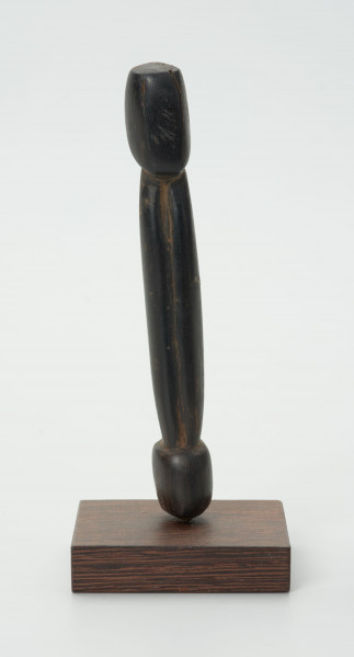 Drewniana figurka postaci z długim tułowiem - Ujęcie z przodu; drewniana figurka postaci ludzkiej przedstawionej schematycznie. Całość wygładzona, zarysowana głowa i długi tułów.