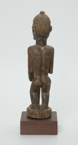 Drewniana figurka kobiety z laską - Ujęcie z tyłu. Drewniana figurka kobiety w pozycji stojącej. W prawej dłoni trzyma laskę. Uszkodzona lewa dłoń. Całość pokryta drobnymi wzorami łącznie z włosami upiętymi w kok.