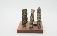rzeźba - Ujęcie z lewego boku; Zestaw składa się z ośmiu małych, drewnianych figurek w pozycji stojącej: siedmiu antropomorficznych i jednej zoomorficznej - przedstawiającej ptaka (dzioborożca calao) na dużym, zaokrąglonym cokoliku. Figurki postaci ludzkich wykonano w sposób schematyczny, dwie mają uniesione do góry ręce. Rzeźby pokryto dość sporą warstwą białej substancji ofiarnej tak grubej, że przesłania ich rzeźbiarską formę.
