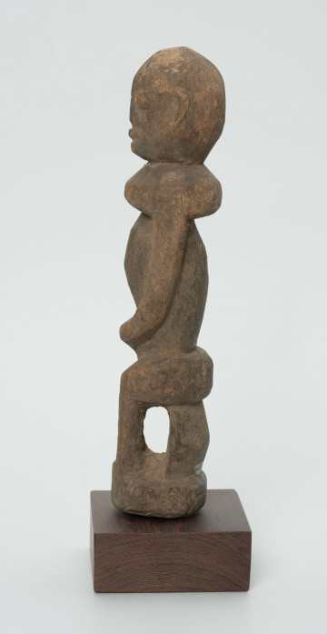 Drewniana figurka postaci siedzącej na stołku - Ujęcie z lewego boku; postać siedząca na stołku. Części ciała i twarz schematyczne, zaznaczone płytkimi nacięciami. Figura statyczna.