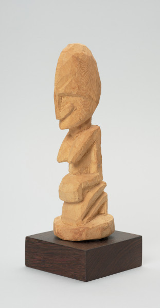 rzeźba - Ujęcie z przodu z lewej strony. Figurka przedstawiająca kobietę siedzącą na podkulonych nogach. Głowa duża, reszta postaci wydobyta płytkimi nacięciami. Forma zgeometryzowana.