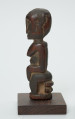 rzeźba - Ujęcie prawej strony rzeźby. Figurka - postać kobiety siedzącej na stołku. O jej płci informują wyraźnie zaznaczone piersi.