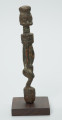 rzeźba - Ujęcie lewej strony rzeźby. Drewniana rzeźba postaci mężczyzny ze zgietymi kolanami. Schematyczne zarysowanie dużej głowy, długiej szyi, na której widoczny jest szeroki naszyjnik. Wąski, długi tułów, długie ręce oparte na kolanach, krótkie nogi i szerokie stopy.