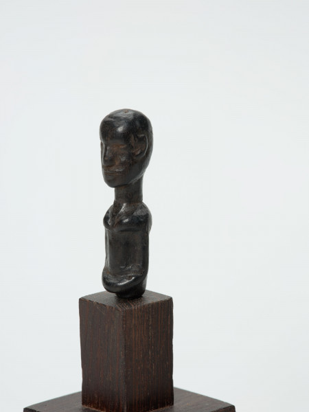 Mała drewniana figurka przedstawiająca popiersie człowieka - Ujęcie ze skosu z lewej; Mała drewniana figurka przedstawiająca popiersie człowieka. Głowa i tułów starannie wygładzone. Postać ujęta schematycznie. Głowa, rysy, piersi, ręce, wydobyte płytkimi nacięciami.