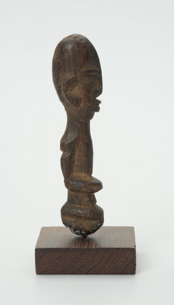 Drewniana figurka z dużą głową i wąskimi oczami - Ujęcie z prawej strony; mała drewniana figurka antropomorficzna. Głowa duża zaokrąglona. Wąskie oczy. Rysy, ręce wydobyte z bryły drewna płytkimi cięciami. Brak nóg.