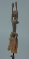 drewniana maska - Ujęcie z tyłu, z prawej strony; Drewniana rzeźbiona maska. Figura postaci ludzkiej z rękoma wzniesionymi do góry. Głowa jajowata, spiczasto zakończona. Zaznaczone uszy. Na wąskim tułowiu wyeksponowane piersi.