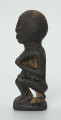 Drewniana figurka kobiety siedzącej na stołku - Ujęcie z boku. Prawy profil figurki łysej kobiety siedzącej na stołku, trzymajacej pierś w lewej dłoni. Powierzchnia figurki chropowata.