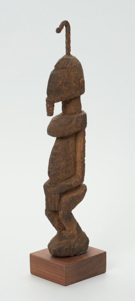 rzeźba - Ujęcie z przodu skosem w lewą stronę. Drewniana rzeźba postaci męskiej. Wąska podłużna twarz, na głowie haczykowata ozdoba. Tułów długi, nogi zgięte w kolanach, ręce oparte na kolanach.
