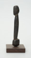 Drewniana figurka postaci z długim tułowiem - Ujęcie z prawego boku; drewniana figurka postaci ludzkiej przedstawionej schematycznie. Całość wygładzona, zarysowana głowa i długi tułów.