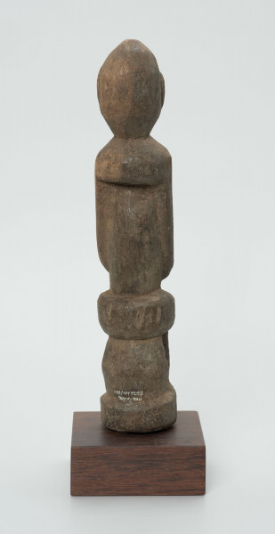 Drewniana figurka postaci siedzącej na stołku - Ujęcie z tyłu; postać siedząca na stołku. Części ciała i twarz schematyczne, zaznaczone płytkimi nacięciami. Figura statyczna.