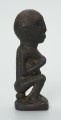 Drewniana figurka kobiety siedzącej na stołku - Ujęcie z boku. Prawy profil figurki łysej kobiety siedzącej na stołku, trzymającej pierś w prawej dłoni. Powierzchnia figurki chropowata.