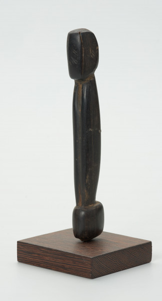 Drewniana figurka postaci z długim tułowiem - Ujęcie z lekkim zwróceniem na lewo; drewniana figurka postaci ludzkiej przedstawionej schematycznie. Całość wygładzona, zarysowana głowa i długi tułów.