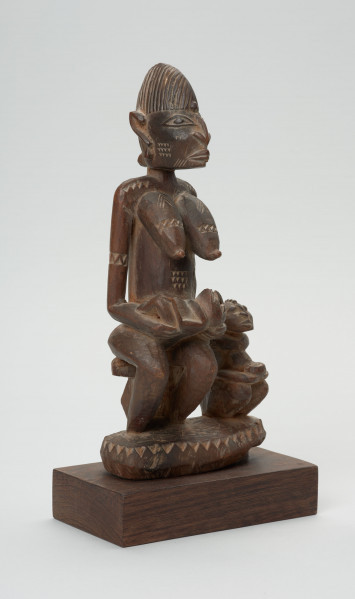 rzeźba - Ujęcie ze skosu z lewej; Figura - kobieta z dziećmi. Rzeźba przedstawia siedzącą kobietę z niemowlakiem na rękach, które opiera na kolanach. Przy niej siedzi drugie dziecko, które kuca. Postać kobiety miejscami rzeźbiona symbolem zygzaka.