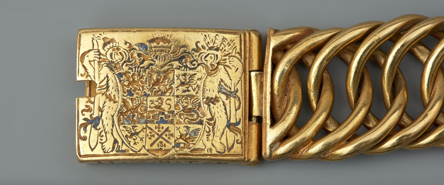 Bransoleta z sarkofagu księcia Franciszka I (1577 - 1620) - Ujęcie lewego zapięcia. Bransoleta wykonana ze skręconych, przenikających się ogniw, w tzw. splocie pancerzowym. Na prostokątnych zapinkach dziewięciopolowy herb Księstwa Pomorskiego trzymany przez dzikich mężów, pierwotnie emaliowany. Na spodzie klamry ryta inskrypcja, pierwotnie emaliowana: V.G.G. / CLARA / G.Z.B.V.L. / H.Z.S.Pō /15.96. Na bocznych ściankach wykonana w tej samej technice inskrypcja: V.G.G. FRANTZ / H.Z.S.Pom.