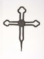 Krzyż żelazny kowalskiej roboty, stanowiący zwieńczenie drewnianego krzyża przydrożnego. Ramiona i trzon wykonane z podwójnych prętów, zakończone elementami dekoracyjnymi o kształcie listka koniczyny.