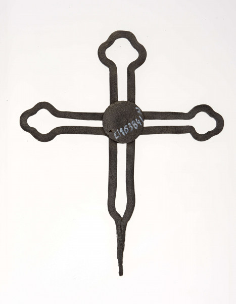 Krzyż żelazny kowalskiej roboty, stanowiący zwieńczenie drewnianego krzyża przydrożnego. Ramiona i trzon wykonane z podwójnych prętów, zakończone elementami dekoracyjnymi o kształcie listka koniczyny.