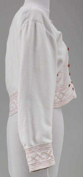 Kaftan kobiecy wykonany z tkaniny bawełnianej (piki), koloru białego. Ozdobiony stebnówką koloru czerwonego i zielonego oraz czerwonymi guzikami. Na lnianej podszewce.