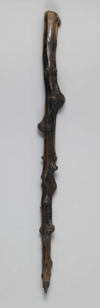 E/1651/ML - Laska drewniana z jednego kawałka drewna; zachowane naturalne wyżłobienia i pozostałości po konarach, tworzące ozdobne guzy. Główka laski spłaszczona, z wyrzeźbioną antropomorficzną twarzą z zaznaczonymi oczami, nosem i ustami. Koniec laski zwężony, zakończony elementem walcowatym oraz ostro zakończonym wielobokiem. Laska politurowana pierwotnie, następnie malowana na całej powierzchni - pozostałości czarnej farby.