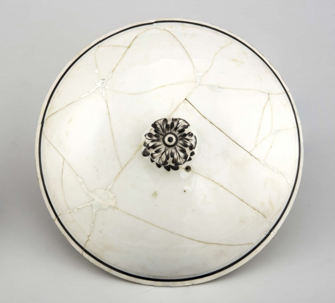 Naczynie stożkowate z uchwytem w postaci szerokiej rurki (służącym do zamocowania drewnianej rączki), pokrywa kopulasta z uchwytem w kształcie rozkwitłej korony kwiatowej. Krawędzie obwiedzione czarnymi paskami, czernią podkreślony modelunek uchwytu. Spód nieszkliwiony.
 