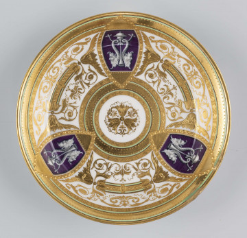 S/CS/1160/ML - Spodek okrągły, konicznie wgłębiony,  na nim bogata dekoracja w wypukłym złocie z ornamentów groteskowych, perełek i wzorów geometrycnych, ułożona w koncentryczne kręgi przedzielone wąskimi paskami jasnej zieleni, z rozetą pośrodku. Na kołnierzu wydzielone 3 migdałowate medaliony, wewnątrz nich mianiatury en grisaille na liliowym tle przedstawiające dwa połączone maszkarony 
  