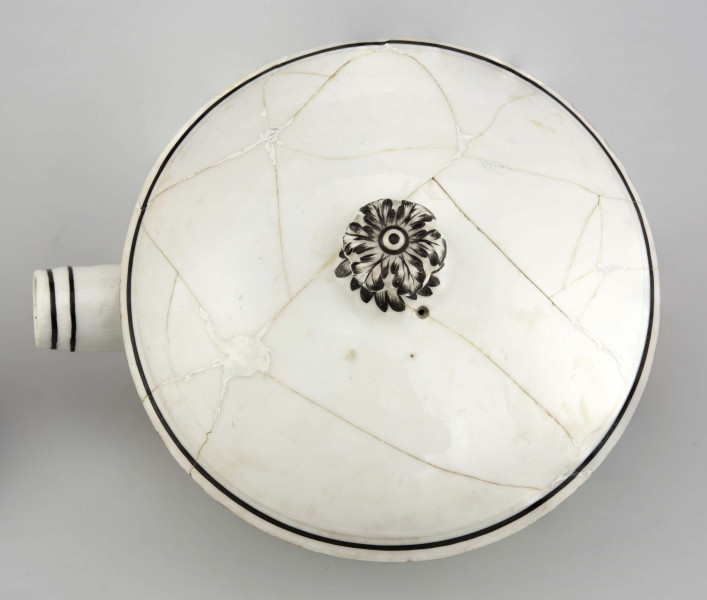 Naczynie stożkowate z uchwytem w postaci szerokiej rurki (służącym do zamocowania drewnianej rączki), pokrywa kopulasta z uchwytem w kształcie rozkwitłej korony kwiatowej. Krawędzie obwiedzione czarnymi paskami, czernią podkreślony modelunek uchwytu. Spód nieszkliwiony.
 