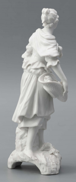 S/CS/1476/ML - Figurka z białej szkliwionej porcelany przedstawiajaca  kobietę stojacąna niskim trójdzielnym cokole. Kobieta ma długie włosy upięte w koczek, ubrana jest w fałdzistą spódnicę przewiązaną poniżej bioder, gorset z baskinką i głębokim dekoltem, bluzkę z bufiastmi rękawami do łokci, na ramionach zawiazaną chustkę, ma bose stopy. Prawą ręką trzyma płaski kosz z owocami oparty na biodrze, w lewej ugiętej ma jabłko. 
  