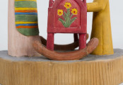 Rzeźba w drewnie lipowym, polichromowana, przedstawiająca postacie matki i dziecka przy kołysce z niemowlęciem. Całość zakomponowana na postumencie wys. 4,4 cm i dł. 22 cm, o kształcie z przodu półokrągłym. Pośrodku usytuowana kołyska na biegunach, wys. 9 cm i dł. 11,2 cm. Kołyska czerwona, ścianki szczytowe ozdobione bukietami składającymi się z trzech wielopłatkowych kwiatów w kolorze żółtym i tulipanowych zielonych liśćmi. W kołysce, w beciku białym, w czerwono-zielone kropki, dziecko, w niebieskiej chustce na głowie. Z lewej strony kołyski postać matki, w długiej jasnoróżowej sukni, w pasie przepasanej fartuchem w żółto-zielono-pomarańczowo-niebieskie poziome pasy. Twarz płaska, nos długi prosty, usta wąskie małe, oczy owalne o podkreślonych białych białkach i czarnych źrenicach, brwi łukowate czarne. Na głowie czerwona chustka zawiązana pod brodą. Spod niej wystają czarne włosy. W rękach trzyma butelkę z mlekiem. Z prawej strony kołyski stoi dziecko w żółtej sukience z niebieskim kołnierzykiem. Twarz dziecka płaska, nos prosty wąski, usta małe, oczy owalne, brwi proste czarne. Włosy czarne, proste sięgające do ramion, zaczesane z grzywką na czole. Ręce wsparte na kołysce. 
