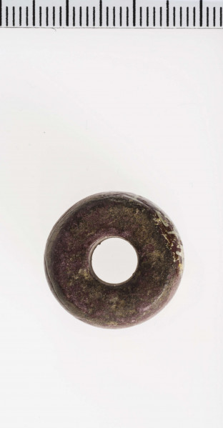 wykonany z łupka owruckiego; kształtu płaskiego walca, bardzo łagodnie półokrągło profilowanego; przy otworze płasko ścięty; otwór dość szeroki, cylindryczny