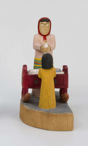 Rzeźba w drewnie lipowym, polichromowana, przedstawiająca postacie matki i dziecka przy kołysce z niemowlęciem. Całość zakomponowana na postumencie wys. 4,4 cm i dł. 22 cm, o kształcie z przodu półokrągłym. Pośrodku usytuowana kołyska na biegunach, wys. 9 cm i dł. 11,2 cm. Kołyska czerwona, ścianki szczytowe ozdobione bukietami składającymi się z trzech wielopłatkowych kwiatów w kolorze żółtym i tulipanowych zielonych liśćmi. W kołysce, w beciku białym, w czerwono-zielone kropki, dziecko, w niebieskiej chustce na głowie. Z lewej strony kołyski postać matki, w długiej jasnoróżowej sukni, w pasie przepasanej fartuchem w żółto-zielono-pomarańczowo-niebieskie poziome pasy. Twarz płaska, nos długi prosty, usta wąskie małe, oczy owalne o podkreślonych białych białkach i czarnych źrenicach, brwi łukowate czarne. Na głowie czerwona chustka zawiązana pod brodą. Spod niej wystają czarne włosy. W rękach trzyma butelkę z mlekiem. Z prawej strony kołyski stoi dziecko w żółtej sukience z niebieskim kołnierzykiem. Twarz dziecka płaska, nos prosty wąski, usta małe, oczy owalne, brwi proste czarne. Włosy czarne, proste sięgające do ramion, zaczesane z grzywką na czole. Ręce wsparte na kołysce. 