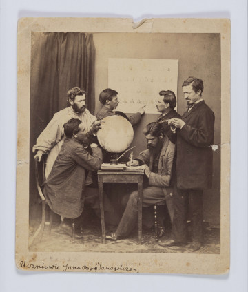 ML/H/F/68 - Jan Bogdanowicz z uczniami na zesłaniu. Fotografia przedstawia sześciu mężczyzn (dwóch siedzących i czterech stojących). Grupa zebrana w pomieszczeniu przy małym kwadratowym stoliku, na którym znajduje się globus, książki, zeszyty i kałamarz z piórem. Jedna z postaci, siedząca po prawej, pisze gęsim piórem. Osoba siedząca naprzeciw wskazuje lewą ręką punkt na globusie. Na ścianie nad stolikiem wisi plansza. Przy niej stoją dwaj mężczyźni - postać po lewej wskazuje coś na planszy prawą dłonią. Nad globusem pochylony jest mężczyzna z brodą w białej długiej koszuli ze stójką (tzw. 
