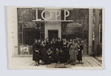 ML/H/F/19/4 - Do fotografii pozuje grupa kilkunastu dziewcząt wraz z ich opiekunką stojącą na skraju grupy po lewej stronie oraz mężczyną w kapeluszu pośrodku. Dziewczęta ubrane są w płaszcze, część ma nakrycia głowy - berety. Stoją na chodniku przed dużą witryną sklepu. Nad wejściem umieszczony duży szydl złożony z białych liter: LOPP. 