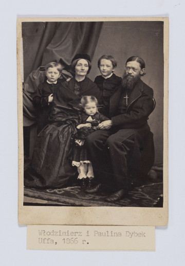 ML/H/F/74 - Fotografia przedstawia rodziców, a pomiędzy nimi stojącą trójkę dzieci. Kobieta w ciemnej sukni okryta szalem, na głowie ma płaski toczek z futra karakułów. Obok na krześle siedzi mężczyzna z dość długimi, gładko zaczesanymi włosami, założonymi za uszy. Ma długą brodę i wąsy. Ubrany w w ciemny garnitur i długi, ciemny płaszcz. Dziewczynka trzyma w ręku lalkę. 
Na odwrociu napis: 