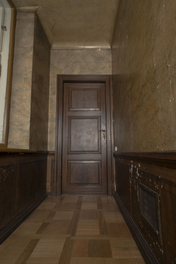 marmoryzowana dekoracja korytarzyka na ścianie E 66M0019, ściany malowane na wzór brązowego marmuru u dołu cokół na wzór boazerii, w tle centralnie zamknięte drewniane drzwi, podłoga drewniana w kwadraty.
