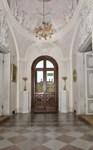 druga część, centralnie drzwi drewniane dwuskrzydłowe prowadzące na dziedziniec, u góry owalna kopuła, po prawej i lewej drzwi wewnętrzne, podłoga wzór dużych rombów, po bokach drzwi dwa marmurowe postumenty z stojącymi na nich złotymi kandelabrami.