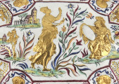 ujęcie z góry - fragment (scena mitologiczna) wieczko. Po lewej stronie tańcząca fama, obok po prawej siedząca postać z tarczą lub lustrem wspartym na kolanach wokół dekoracja roślinna, w lewym rogu zamek.