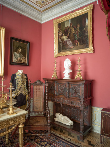 narożnik południowo-wschodni, różowe ściany po prawej  stoi drewniana komoda, na ścianie duży obraz, po lewej manekin w stroju z epoki, po środku krzesło.