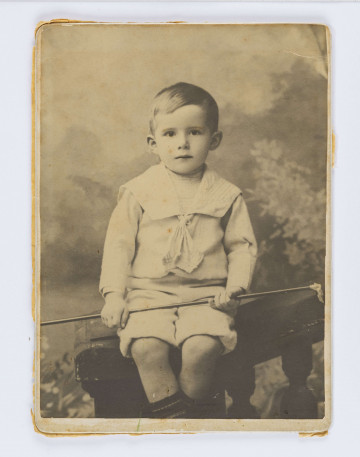 Dziecko w wieku ok. 4 lat. Siedzi na ławeczce, w kożuszku. Na głowie czapka owinięta szalikiem, w prawym ręku trzyma długą laseczkę.