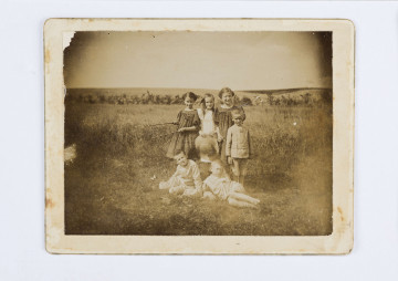 Fotografia Adam Żeromskiego w grupie dzieci na tle pola. Na ziemi siedzi dwóch chłopców, za nimi stoją trzy dziewczynki i jeden chłopiec.