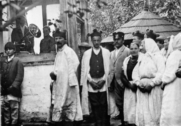Fotografia czarno-biała przedstawia grupę w strojach ludowych. Mężczyźni i kobiety stoją blisko zabudowań wiejskich. Po lewej stronie, wewnątrz przybudówki znajdują się odświętnie ubrani mężczyzna i kobieta z instrumentem muzycznym. 