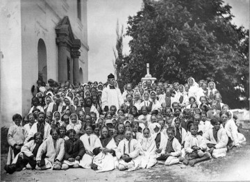 Fotografia czarno-biała przedstawia grupę kilkudziesięciu dzieci w strojach ludowych oraz księdza. Po lewej stronie widoczny fragment wejścia do kościoła, po prawej stronie rozłożyste drzewo.  