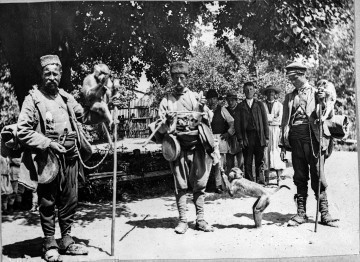 Fotografia plenerowa czarno-biała przedstawia grupę mężczyzn. Trzy postacie na pierwszym planie to kuglarze, każdy z nich ukazany wraz z małpą. Na drugim planie czterech mężczyzn w strojach ludowych. 