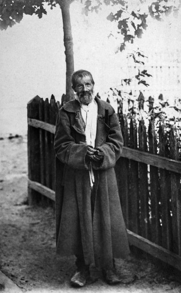 Fotografia czarno-biała przedstawia starszego mężczyznę w długim płaszczu, stojącego przy drewnianym płocie.  