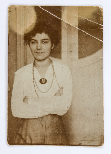 Fotografia w formie stojącego prostokąta, przedstawiająca  Katarzynę Czechowiczówną (3/4 ujętej postaci) na tle białych, oszklonych drzwi. Katarzyna stoi nieco po lewej stronie, z załozonymi rękami. Ubrana jest w białą bluzkę i spódnicę. Na szyi ma drobne korale, do bluzki przypięta ciemna broszka. Włosy ciemne, kręcone, upięte z tyłu, ciemne, grube brwi, na twarzy lekki uśmiech. Fotografia monochromatyczna, sepia. Powierzchnia papieru gładka, lekko świecąca. Nad głową postaci dwie białe plamy. Na odwrocie, w górnej części ślady po kleju, nizej numer inwentarza napisany ołówkiem.