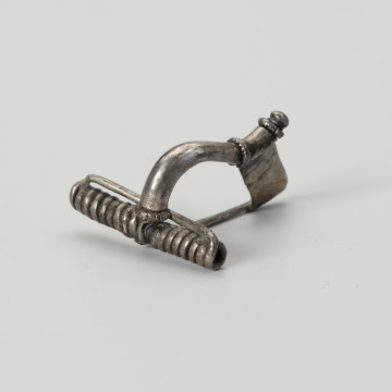 Fibula - ujęcie ze skosu; Fibula - zapinka srebrna posiada niewielki guzek na zakończeniu kabłąka, który dodatkowo jest zdobiony dwoma karbowanymi pierścieniami znajdującymi się u nasady nóżki. Podobny trzeci pierścień umiejscowiony jest na kabłąku bezpośrednio nad sprężynką.