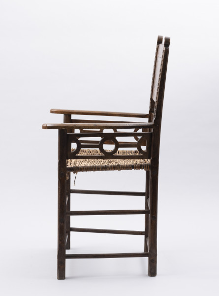 Fotel - ujęcie z boku; Fotel - krzesło z podłokietnikami. Mebel o konstrukcji ramowej z pionowo wznoszącym się oparciem. Nogi proste o przekroju prostokątnym, łączone dwiema toczonymi podpórkami, przednie z delikatnym wzorem geometrycznym wykonanym techniką snycerską. Siedzisko proste, wyplatane wikliną. Pomiędzy siedziskiem, dolną częścią oparcia i podłokietnikami ażurowo ozdobnie wycięta listwa ( motyw koła) będąca zarazem elementem konstrukcji. W ramę, w jej górnej części wpuszczona deska.